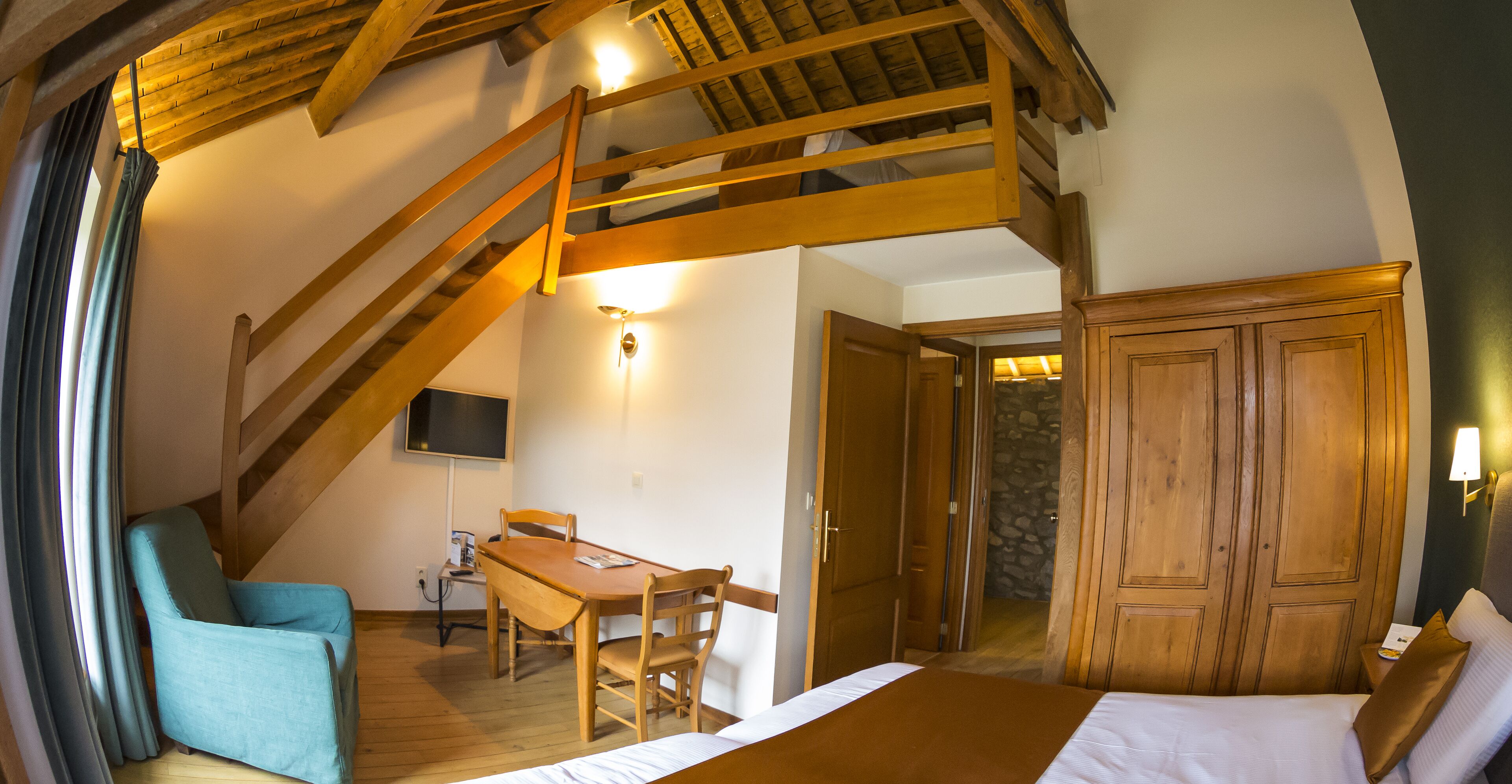A room with a bed, a desk, and a chair. There is a wooden staircase leading to an upper level.