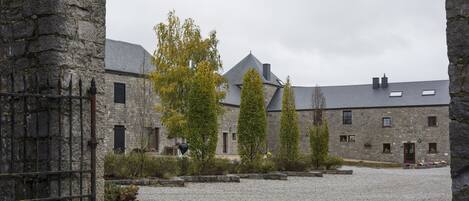 Un bâtiment en pierre avec une cour recouverte de gravier, un portail en métal et une rangée d’arbres.