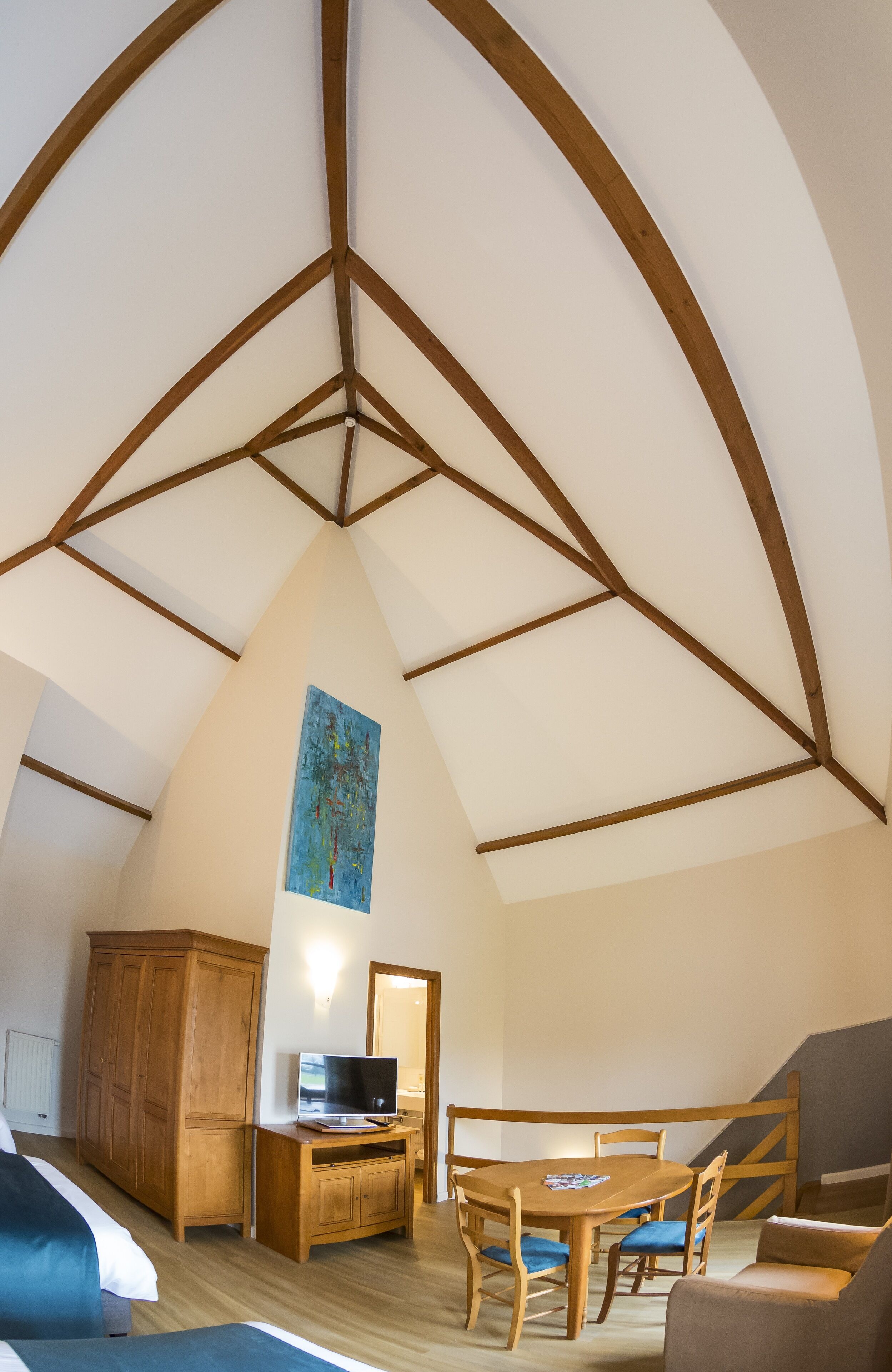 A room with a sloped ceiling, wooden beams, a dining table with chairs, a television on a wooden stand, and a wardrobe.