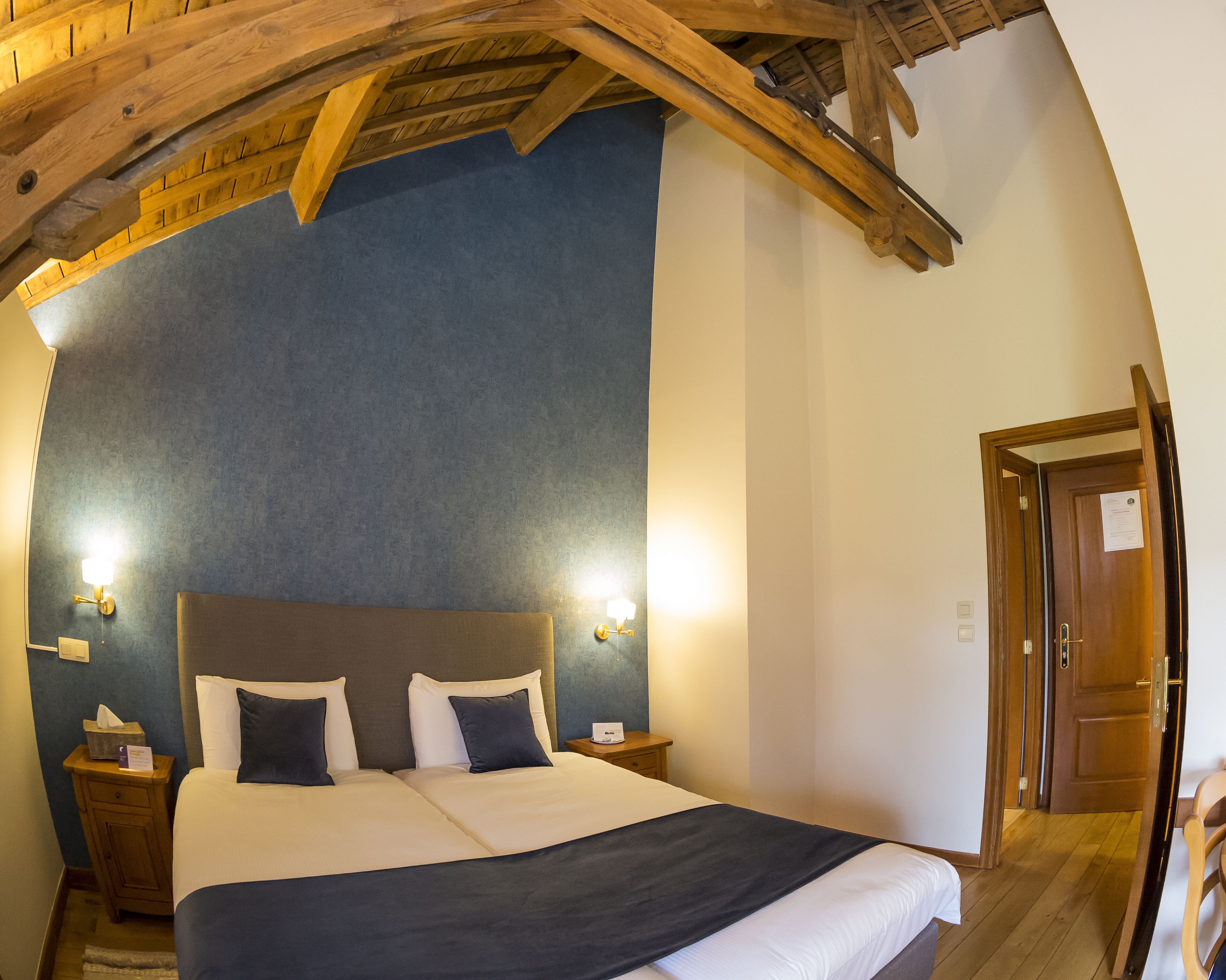 A hotel room with a large bed, wooden ceiling beams, two bedside lamps, and a wooden door.