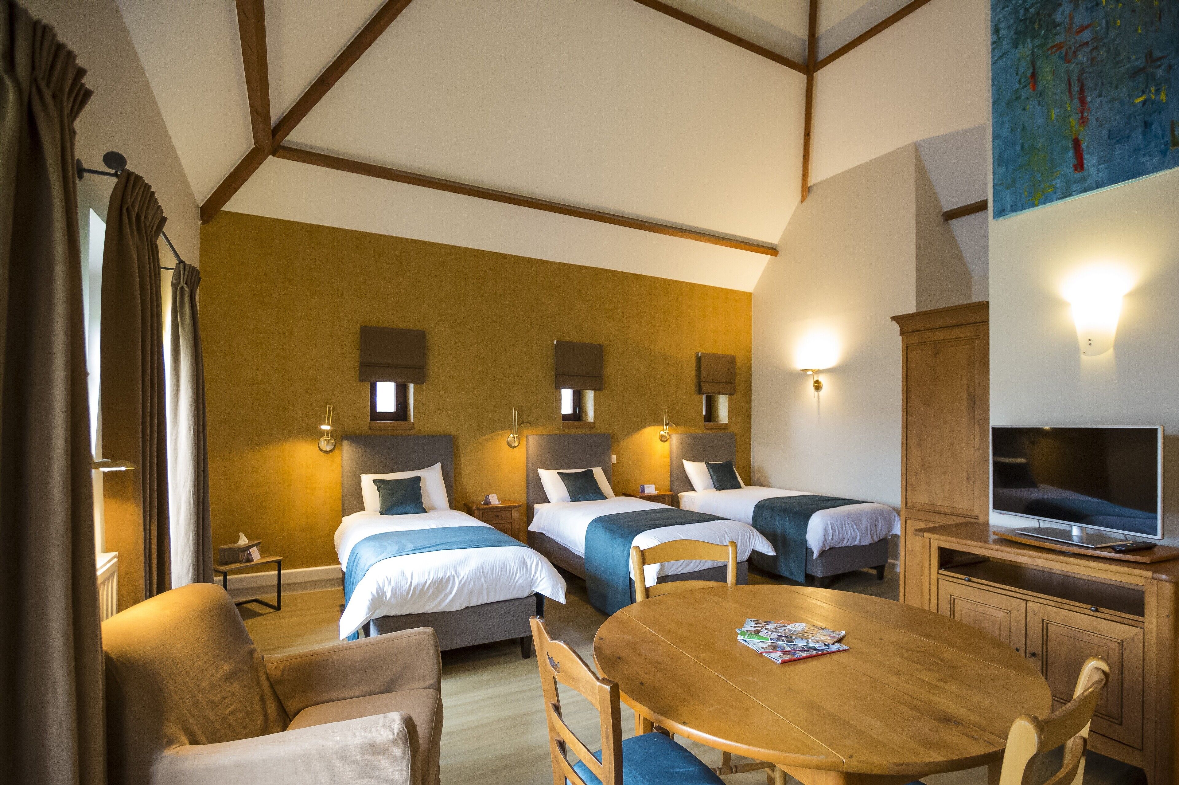 A hotel room with three single beds, a wooden dining table, a television, and a painting on the wall.
