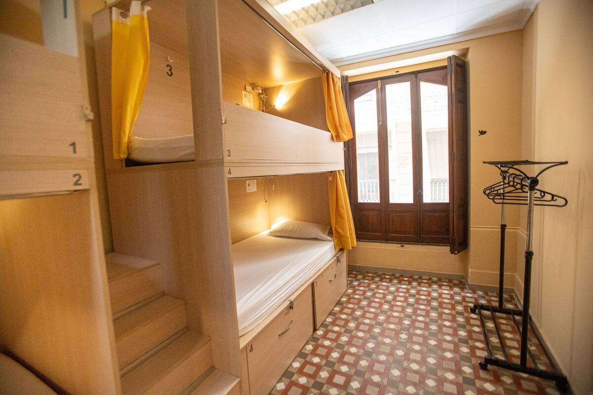 Un dormitorio con literas, taquillas numeradas, una ventana con cortinas y un tendedero.