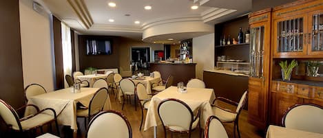 Un restaurante con varias mesas y sillas, una zona de bar con botellas, y una pantalla de televisión en la pared.