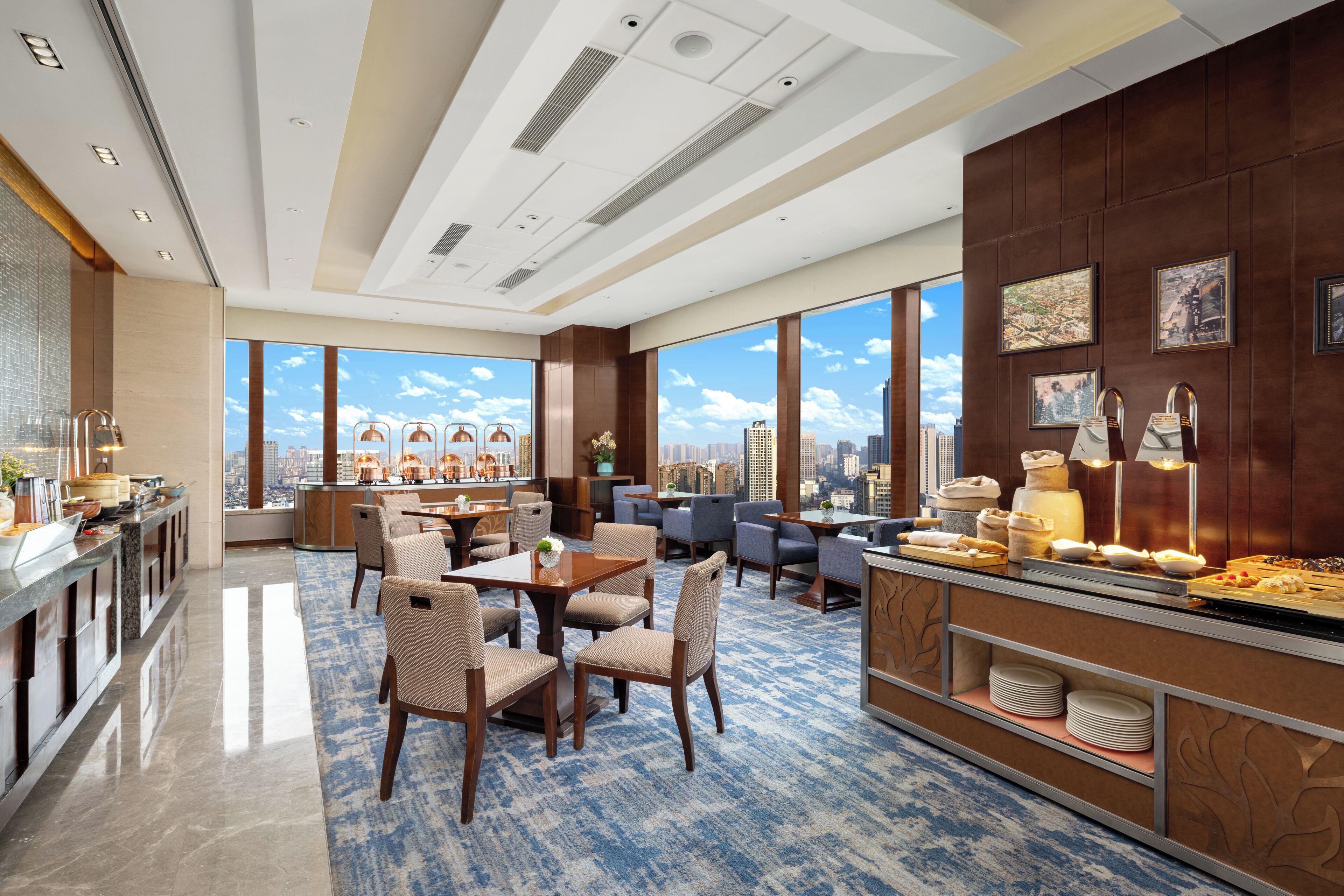 A modern hotel lobby with a dining area, large windows offering city views, and a buffet with various food items.