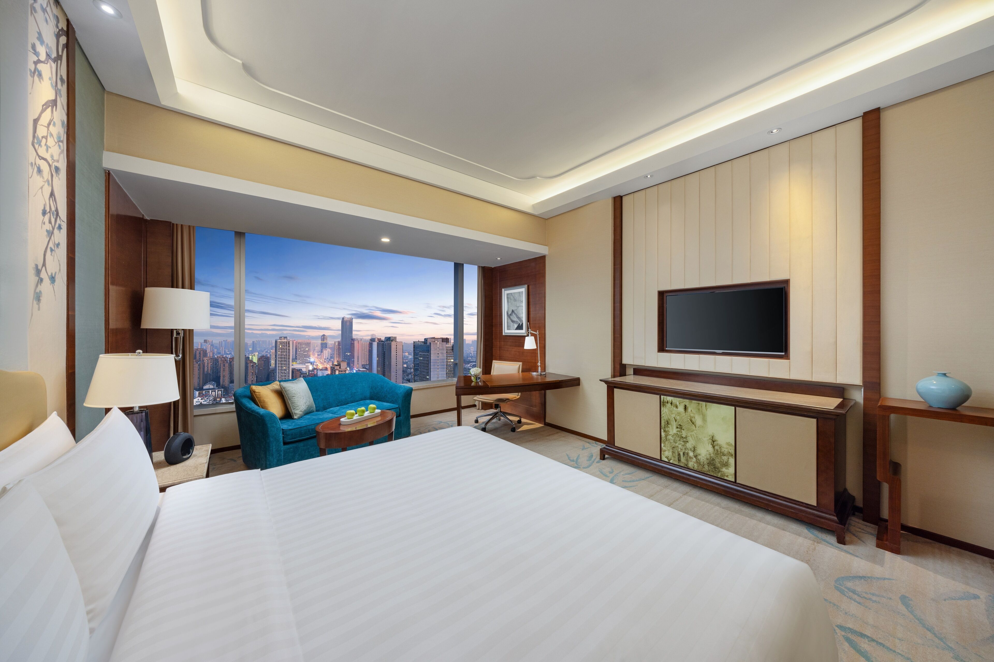 A hotel room with a large bed, a desk, a chair, a TV, and a view of the cityscape.