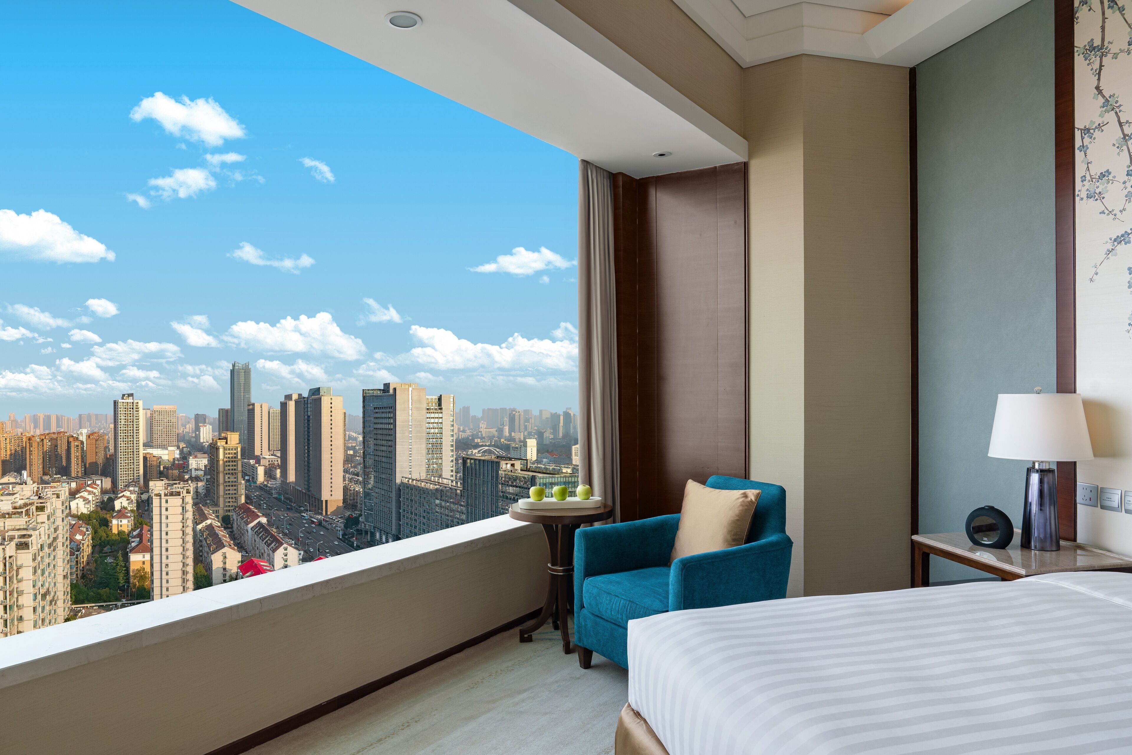 Une chambre d’hôtel avec vue sur la ville, un lit, un fauteuil bleu, une petite table avec des pommes et une lampe.