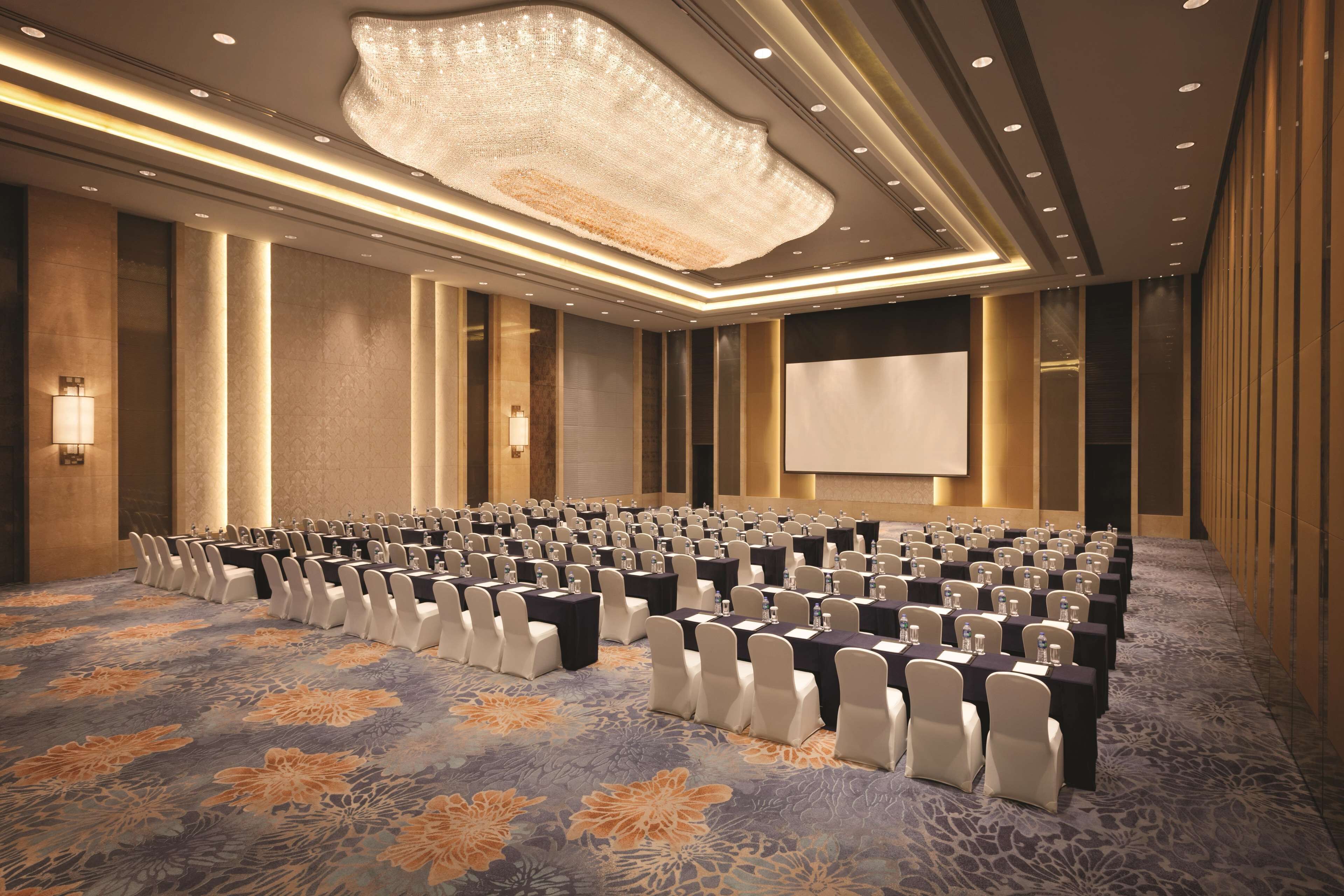 A conference room with rows of tables and chairs, a large screen, and a chandelier.