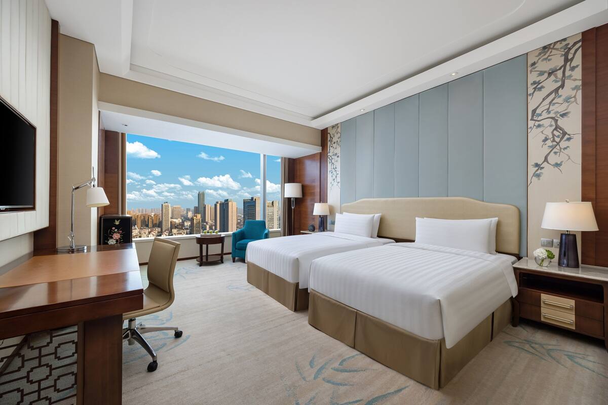 A hotel room with two beds, a desk, a chair, a TV, and a large window with a city view.