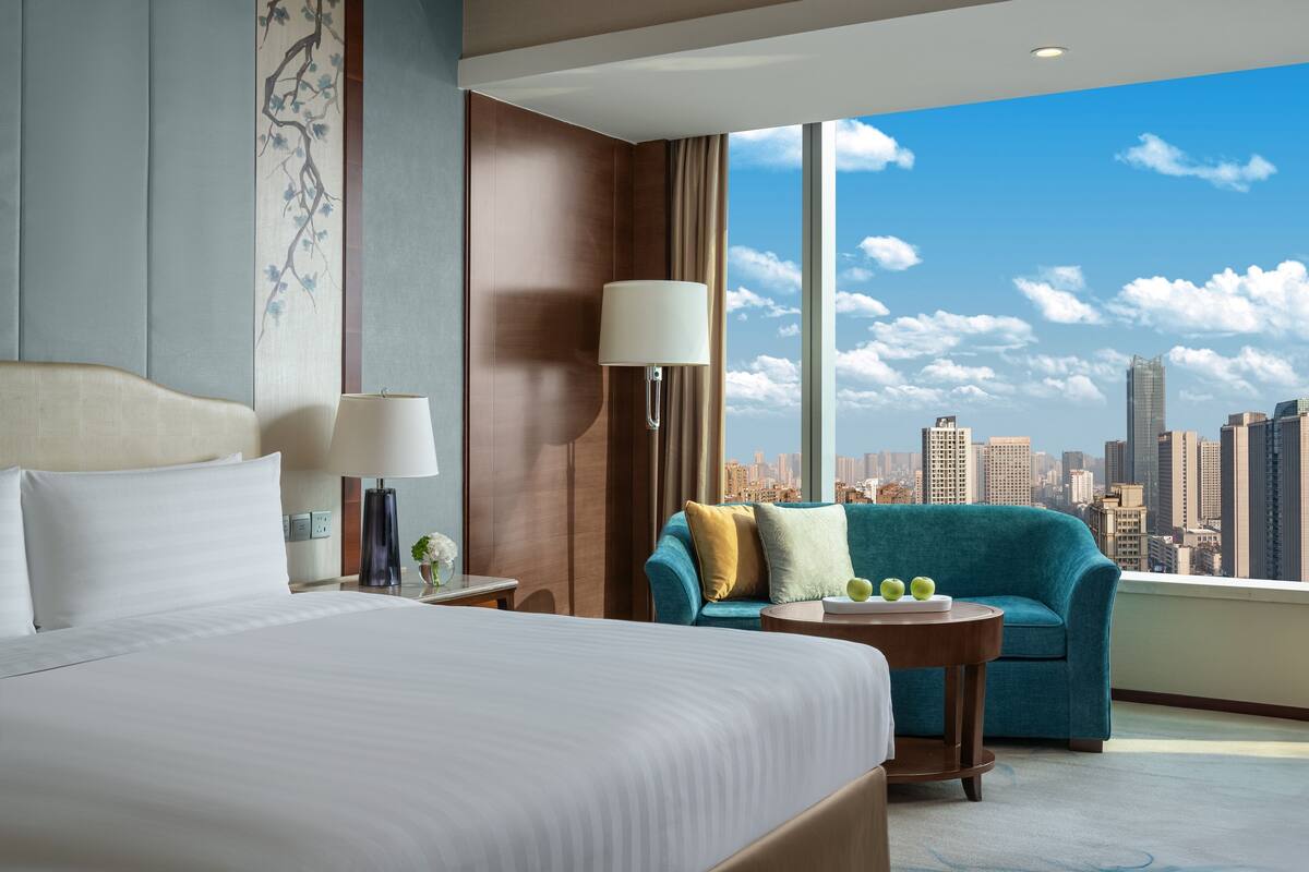 A hotel room with a large bed, a sofa, a small table, and a view of the city skyline.