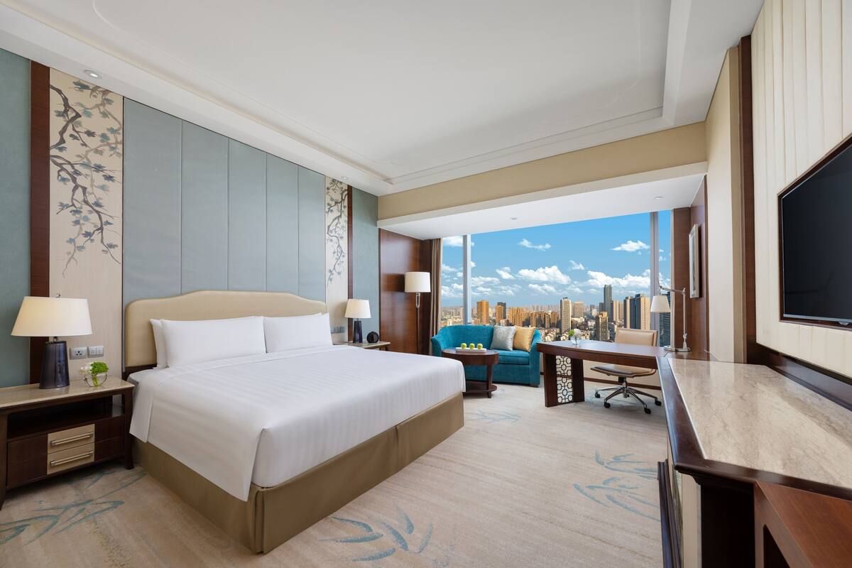 A hotel room with a large bed, a desk, a sofa, and a TV. The room has a city view.