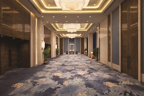 A hotel corridor with a patterned carpet, chandeliers, and potted plants.