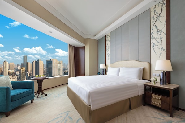 Habitación de hotel con una cama grande, vistas a la ciudad, un sillón azul y una mesita de noche con una lámpara.