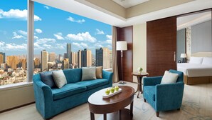 A modern hotel room with a large window offering a city view, a blue sofa, a small table with apples, and a separate armchair. - Shangri-La Hefei (Hefei)