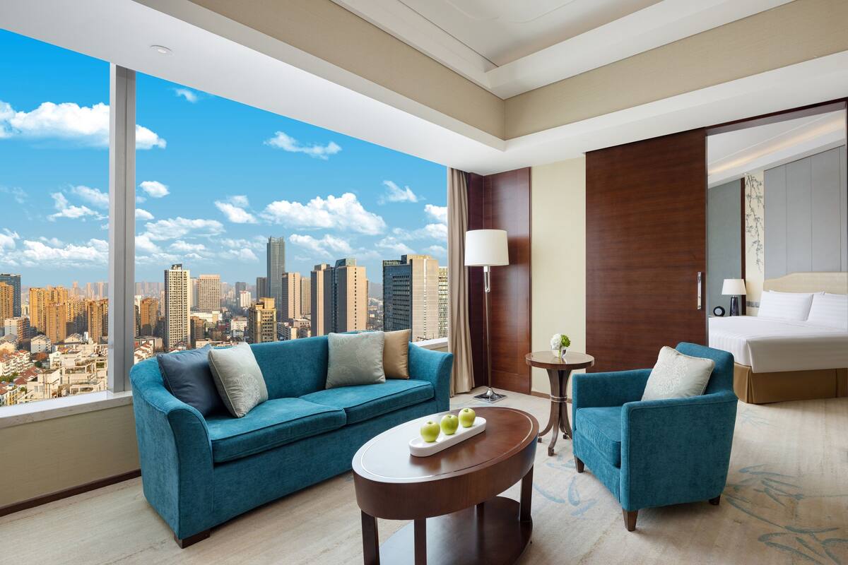 A modern hotel room with a large window offering a city view, a blue sofa, a small table with apples, and a separate armchair.