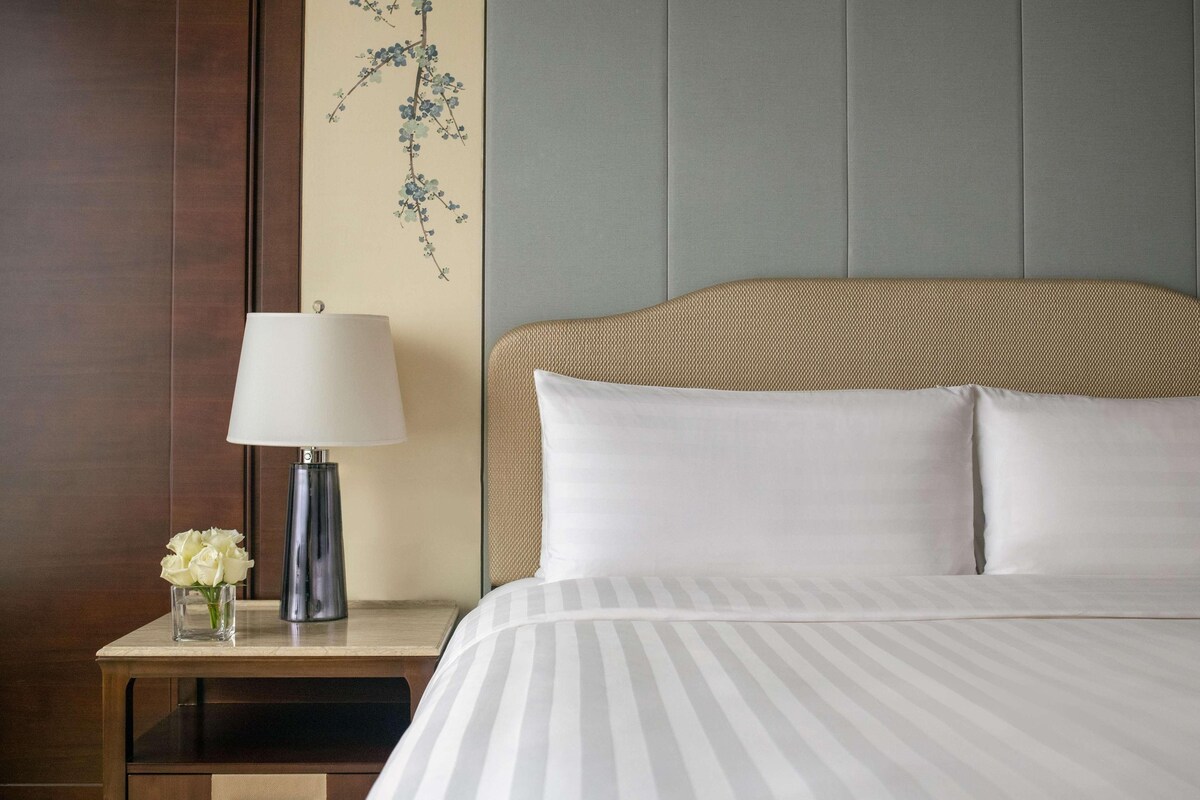 A neatly made bed with white and gray striped bedding, a wooden nightstand with a lamp and a vase of white flowers, and a wall with a floral tapestry.