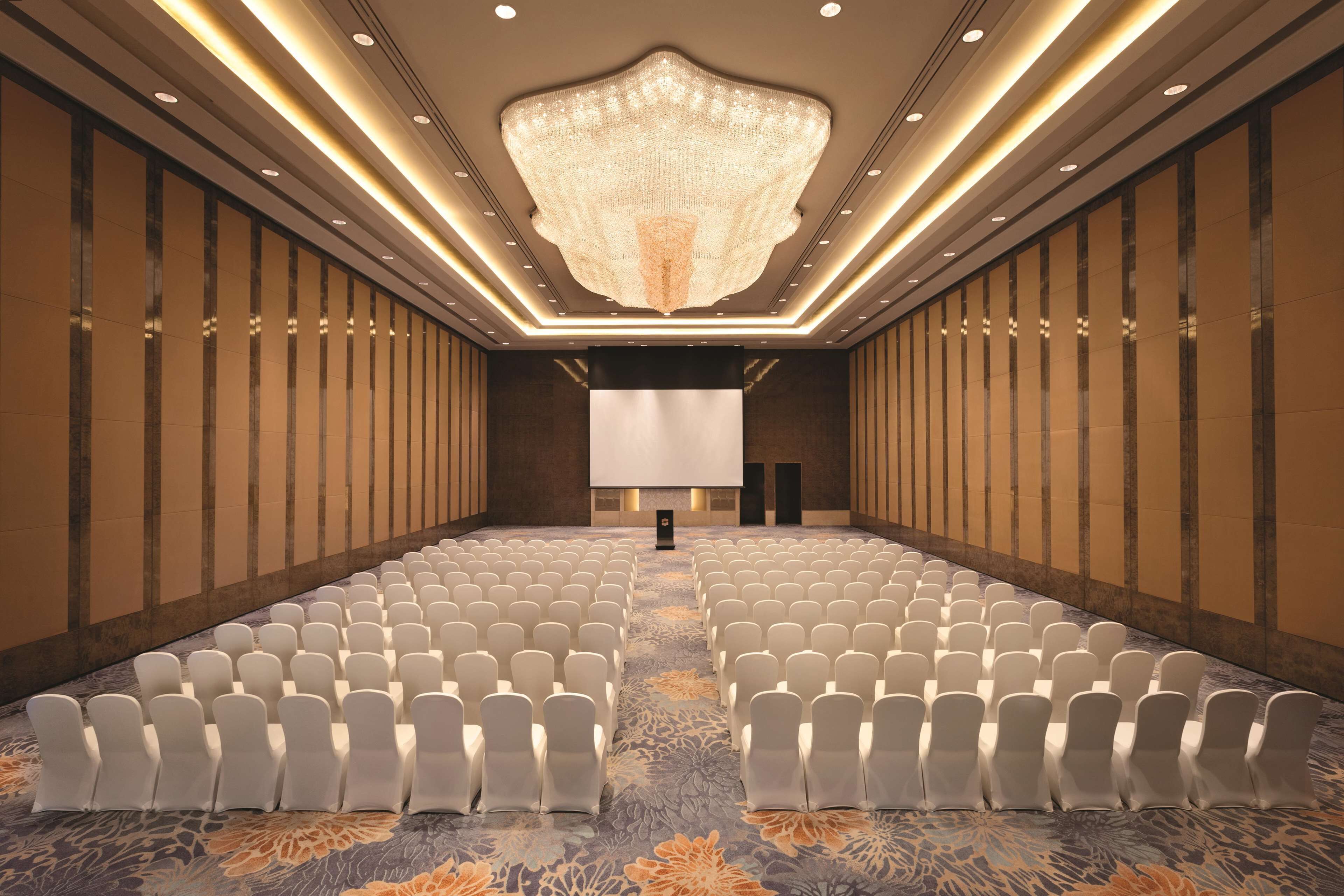 A conference room with rows of white chairs, a large screen at the front, and a chandelier.
