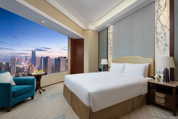 A hotel room with a large bed, a blue armchair, a nightstand with a lamp, and a view of a cityscape through a large window. - Shangri-La Hefei (Hefei)