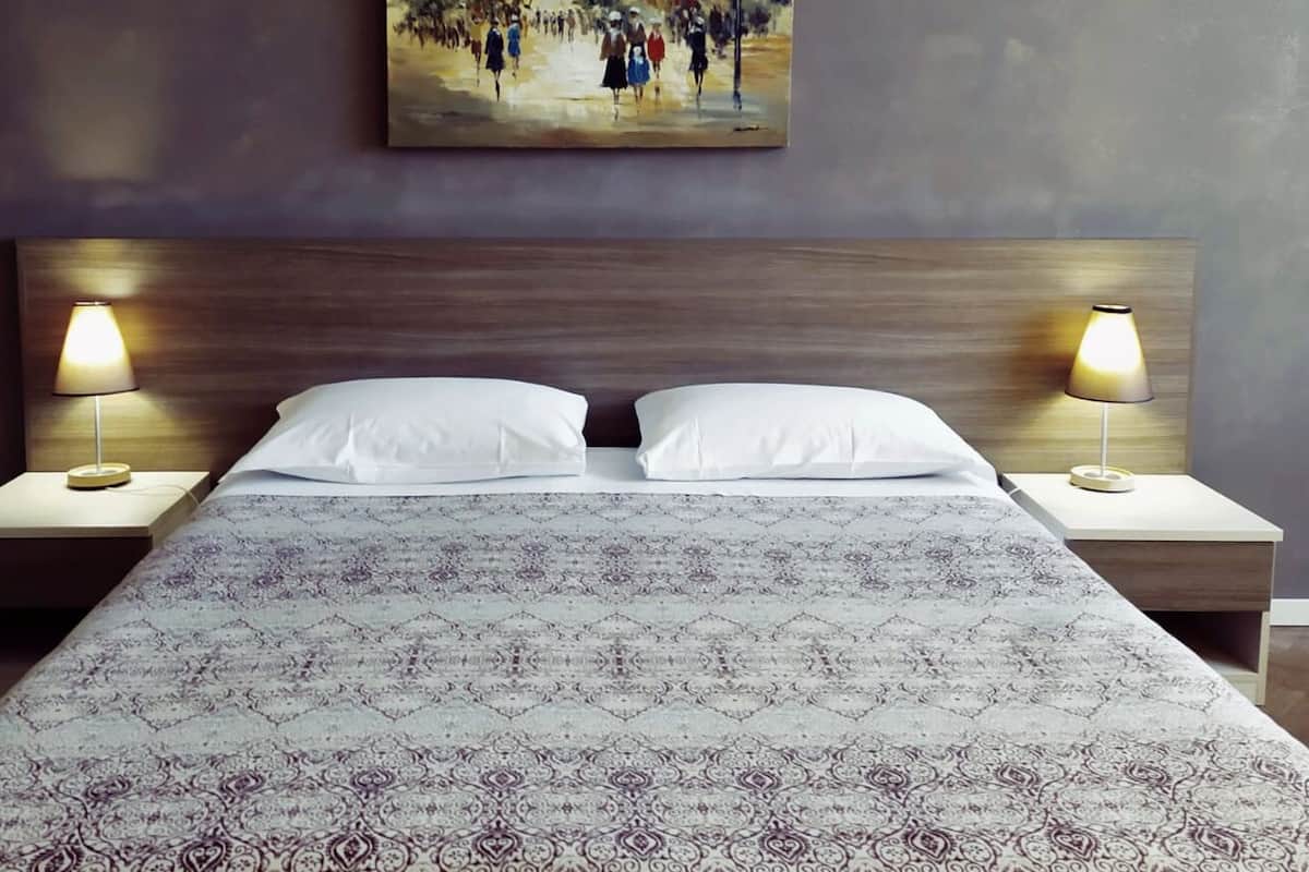 Quarto de hotel com uma cama grande, duas luminárias laterais e um quadro na parede.