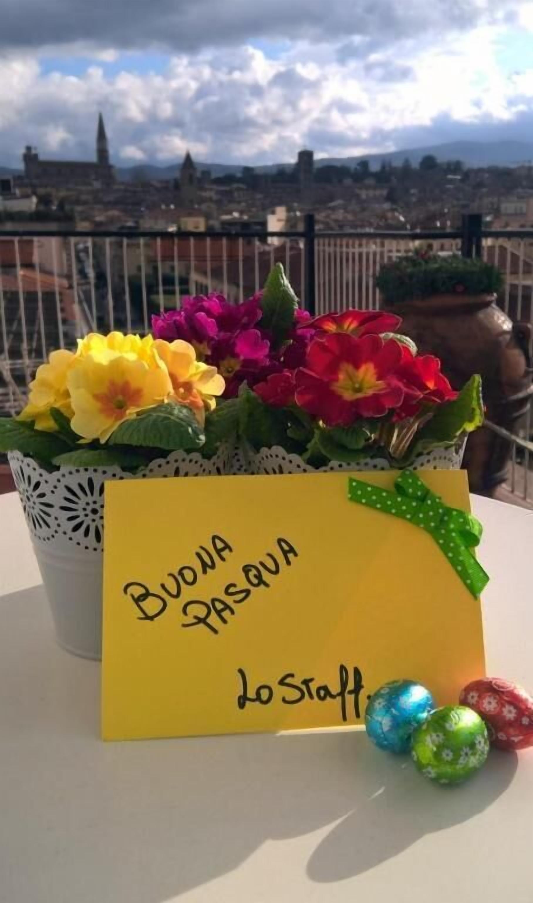 A yellow card with "Buona Pasqua Lo Staff" written on it, placed on a table with a view of a cityscape and a pot of flowers.