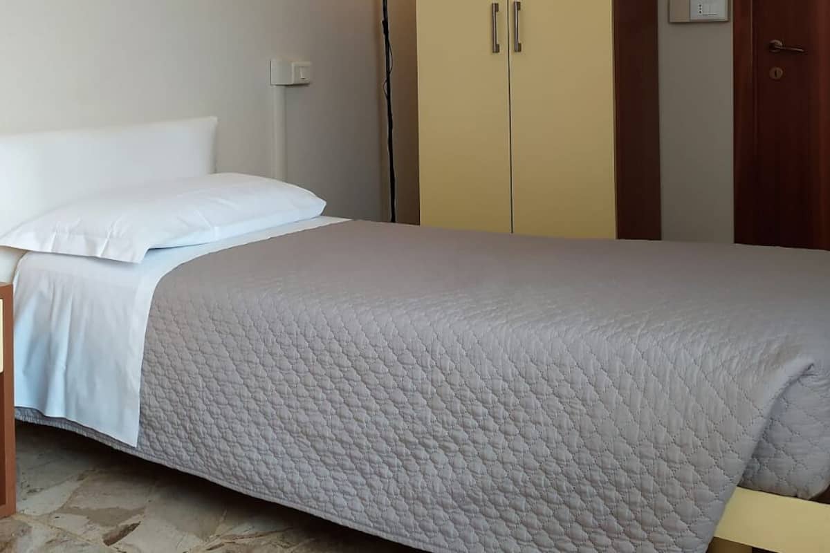 Uma cama de solteiro com cobertura acolchoada, uma mesa de cabeceira, um abajur e um guarda-roupa num quarto com chão em mosaico.