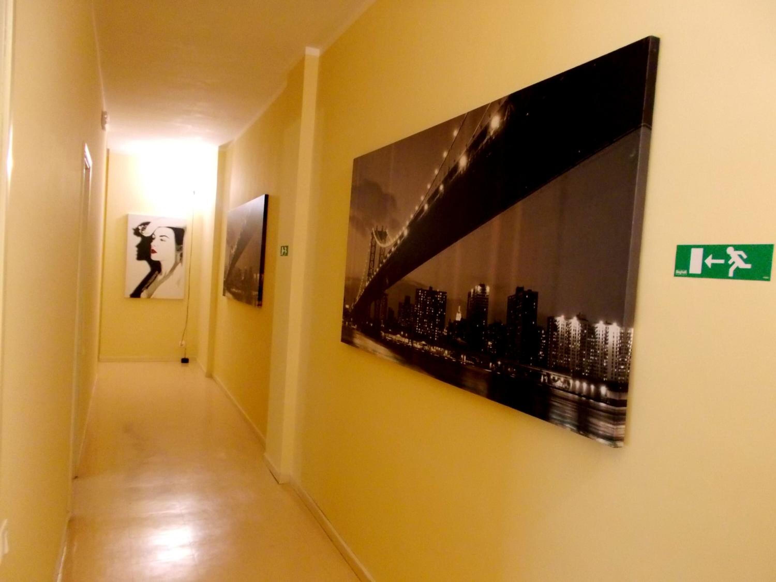A hallway with a large cityscape painting, a smaller portrait, and an emergency exit sign.
