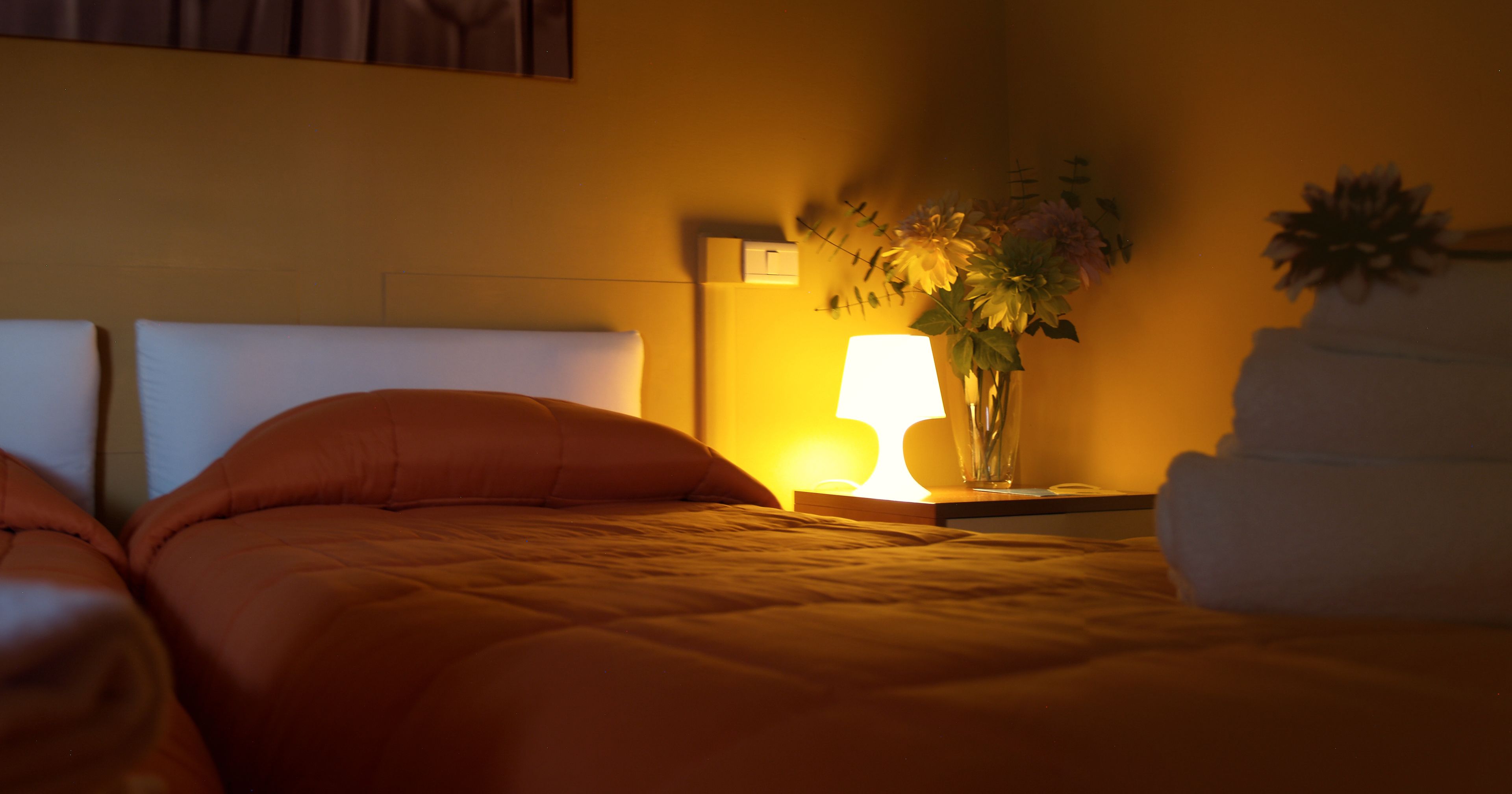 Cama bem arrumada com manta laranja, luminária de cabeceira e um vaso com flores sobre a mesa de apoio.