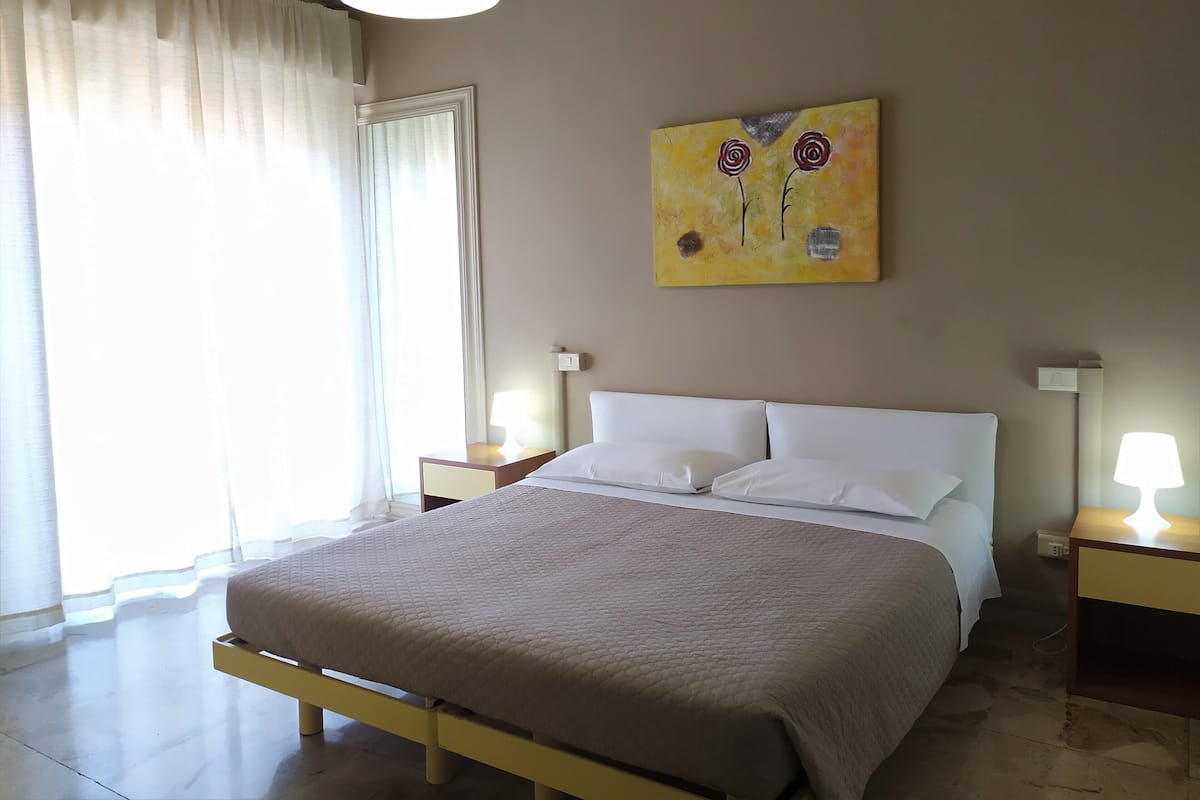Quarto de hotel com cama, duas mesas de cabeceira, um quadro na parede e uma janela com cortinas.