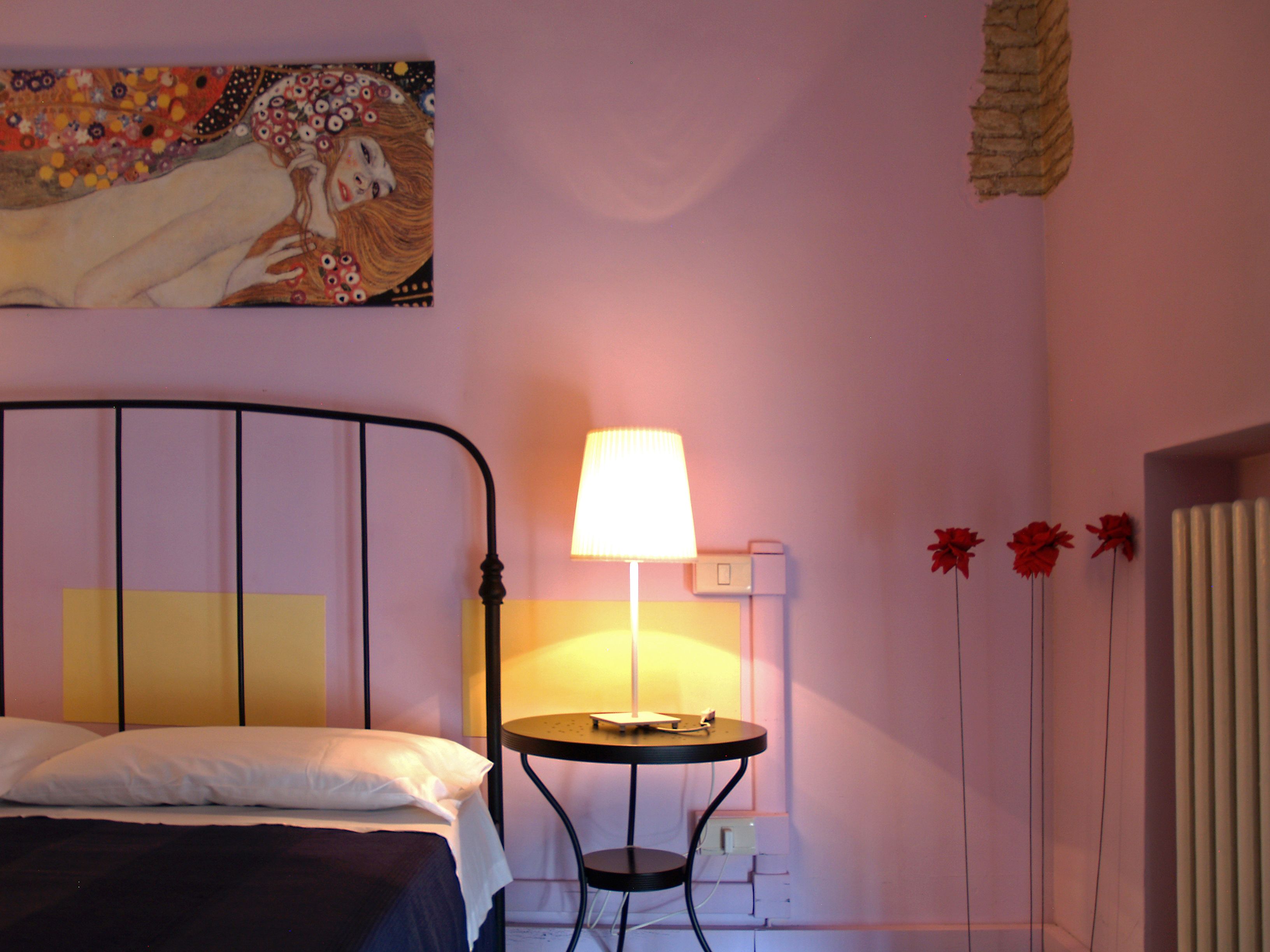 A bedroom with a bed, a bedside table with a lamp, a wall-mounted power outlet, and a decorative flower arrangement.