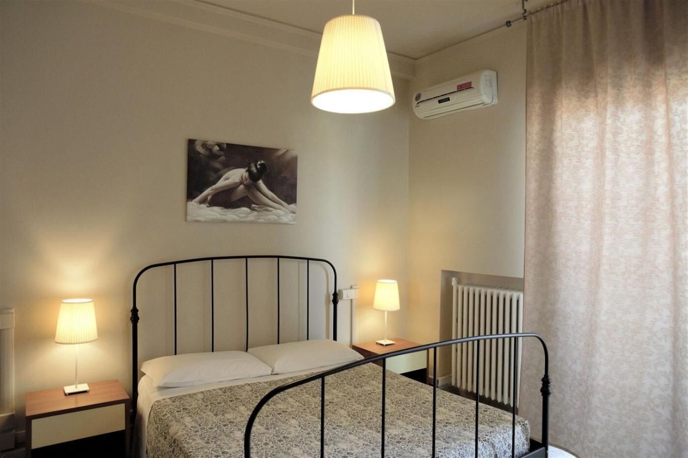 Un dormitorio con una cama, dos lámparas de noche, un radiador, un equipo de aire acondicionado y un cuadro en la pared.