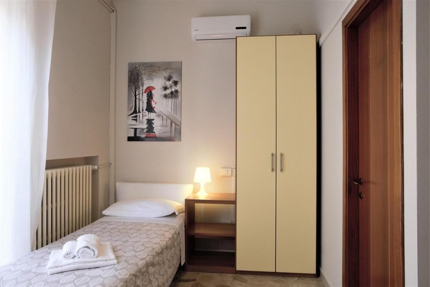 A small, neatly arranged bedroom with a bed, a bedside lamp, a wardrobe, and a radiator.