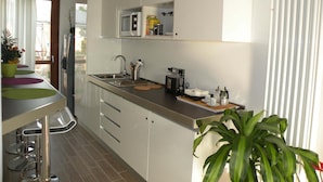 A modern kitchen with white cabinets, a stainless steel sink, a microwave, and a coffee machine. There are potted plants on the countertops and a window with curtains. - Cameracaffè sul Lago (Arezzo)
