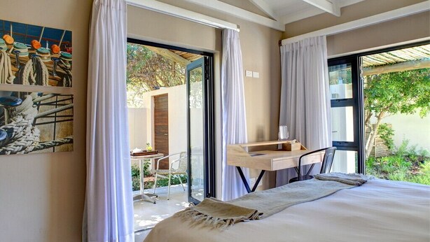 A bedroom with a large bed, a desk, and a chair. There are curtains on the windows, and a painting is on the wall. - Scallop Guest Lodge (Plettenberg Bay)