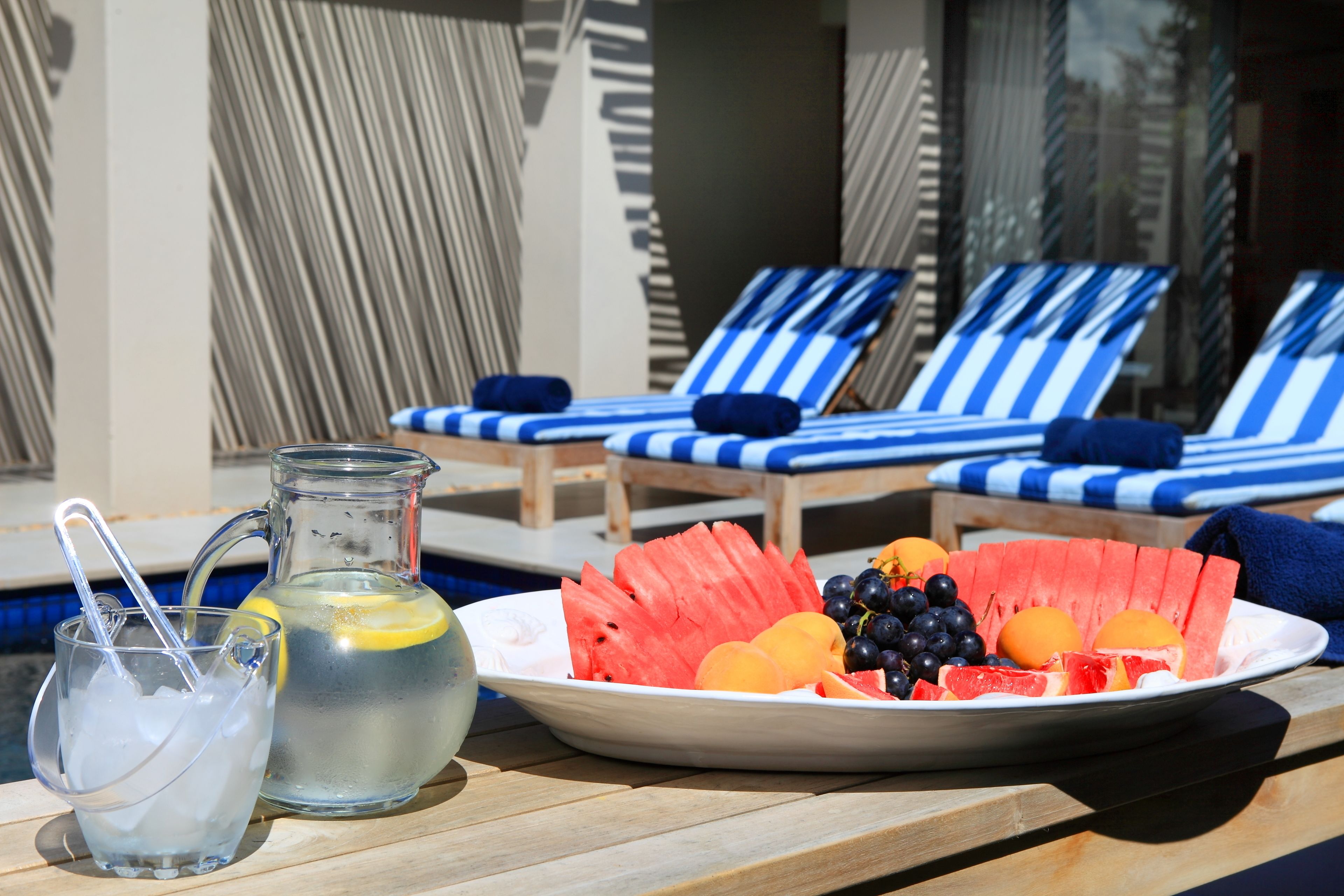 Une table placée au bord de la piscine, sur laquelle on trouve une assiette de pastèque, de raisins et d’oranges, une carafe de limonade et un verre d’eau glacée.