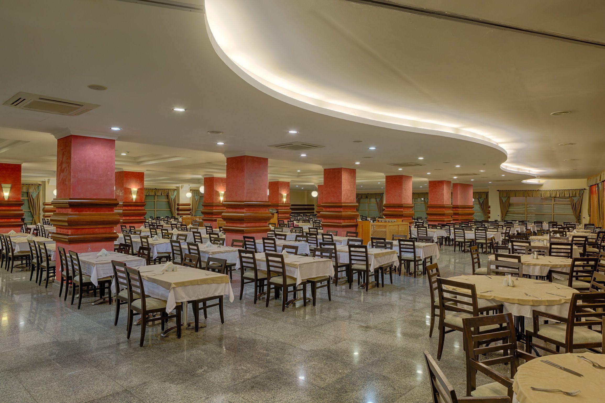 Un amplio comedor con numerosas mesas y sillas, techos altos con iluminación empotrada y pilares rojos.