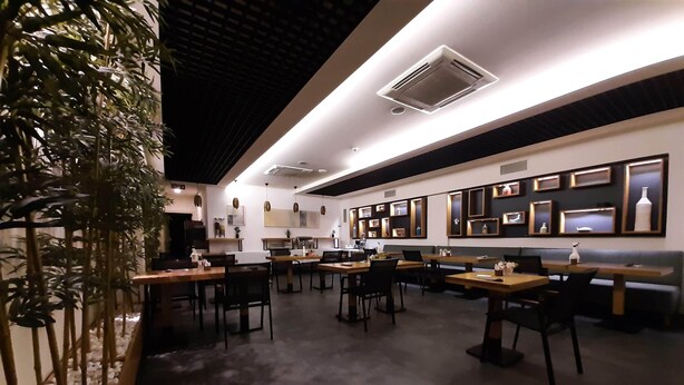 Un restaurant moderne avec des tables et des chaises en bois, un plafond haut doté d’un éclairage encastré et des étagères décoratives fixées au mur.