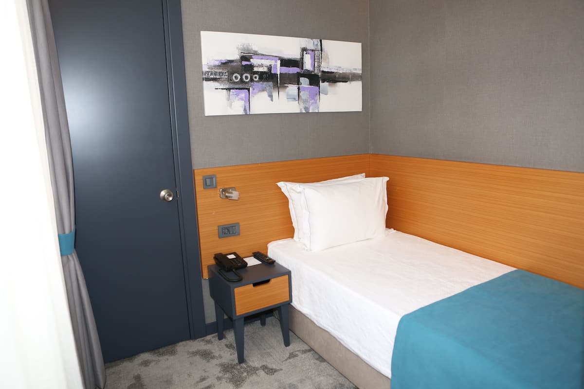 A hotel room with a wooden headboard, a single bed, a nightstand with a phone and remote, a gray wall with a painting, and a door with a curtain.