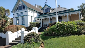A two-story building with a white facade and blue trim, labeled "Yellowwood Lodge." It features a covered porch with white lattice detailing and a small garden area with various plants and flowers. - Yellowwood Lodge - Adults Only (Knysna)