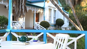 A white and blue house with a porch, a table with a teapot and a bowl, and a chair. - Yellowwood Lodge - Adults Only (Knysna)