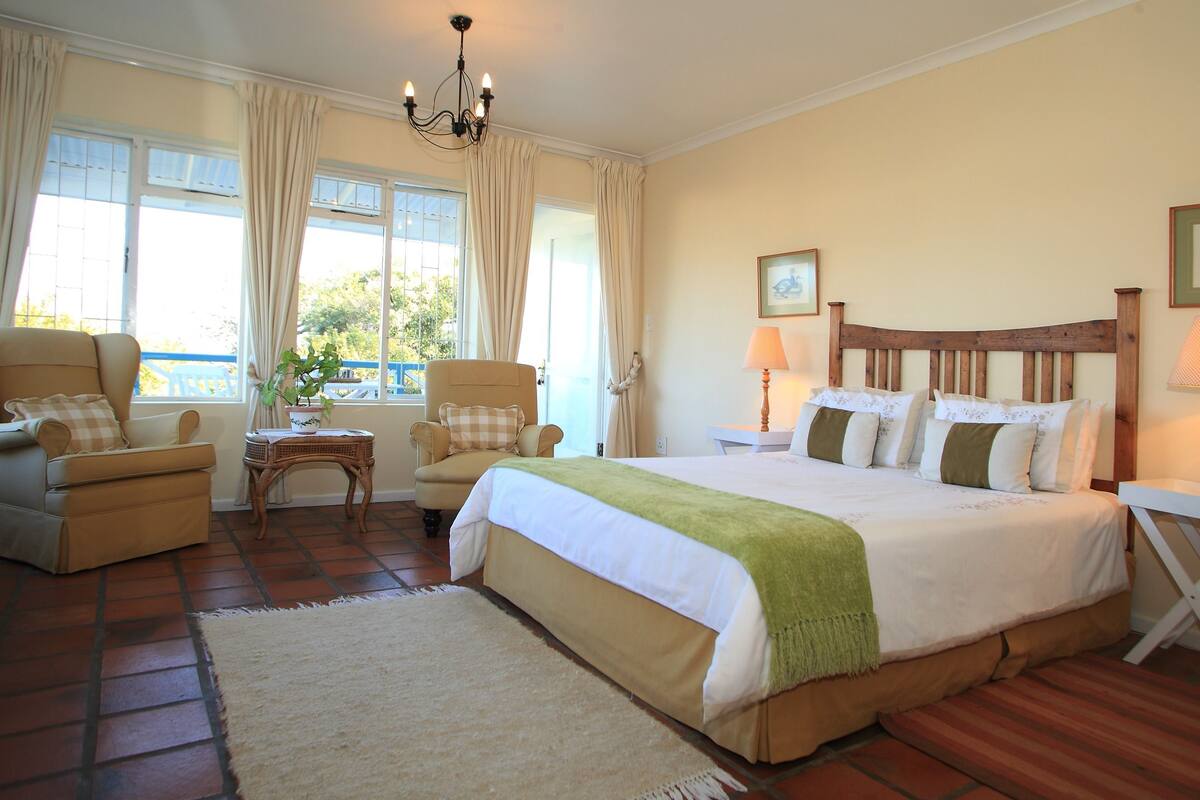 A bedroom with a large bed, two bedside tables, a chair, a small table, and a view of the outdoors.