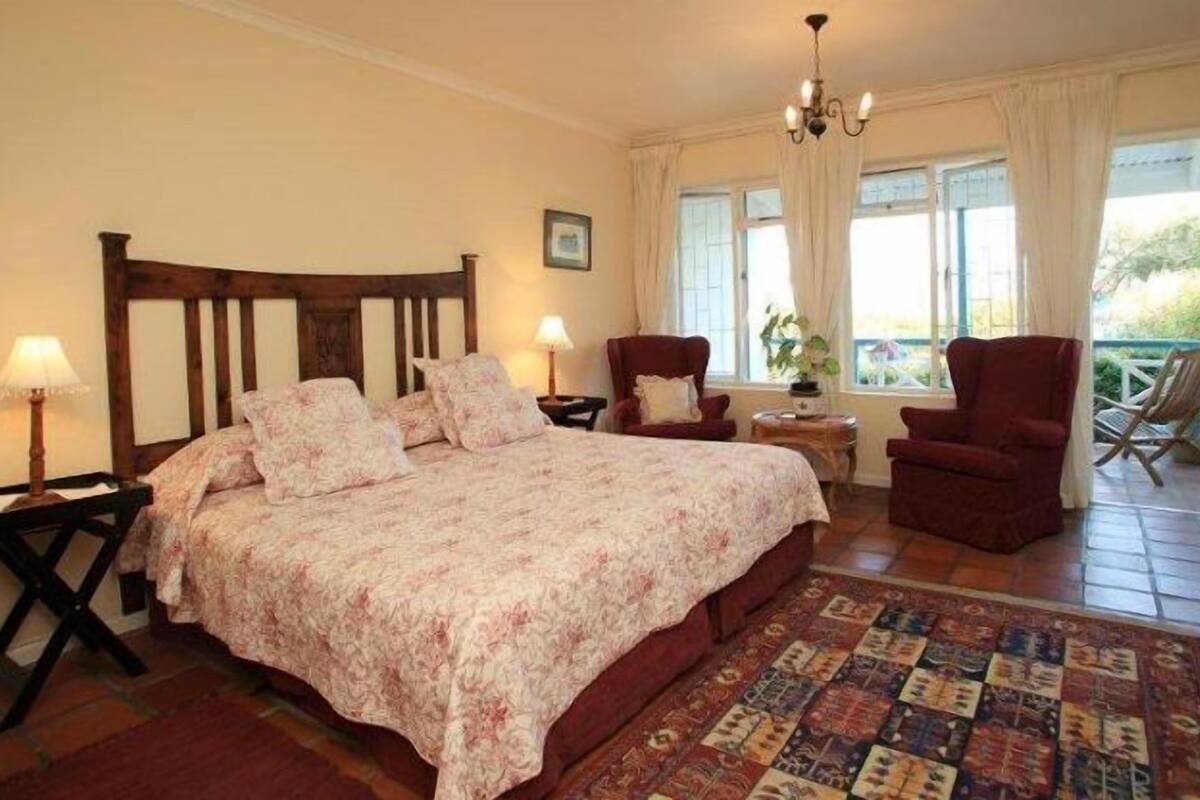 A bedroom with a wooden bed, two bedside tables with lamps, a red armchair, a view of the outdoors through a window, and a patterned rug on the floor.