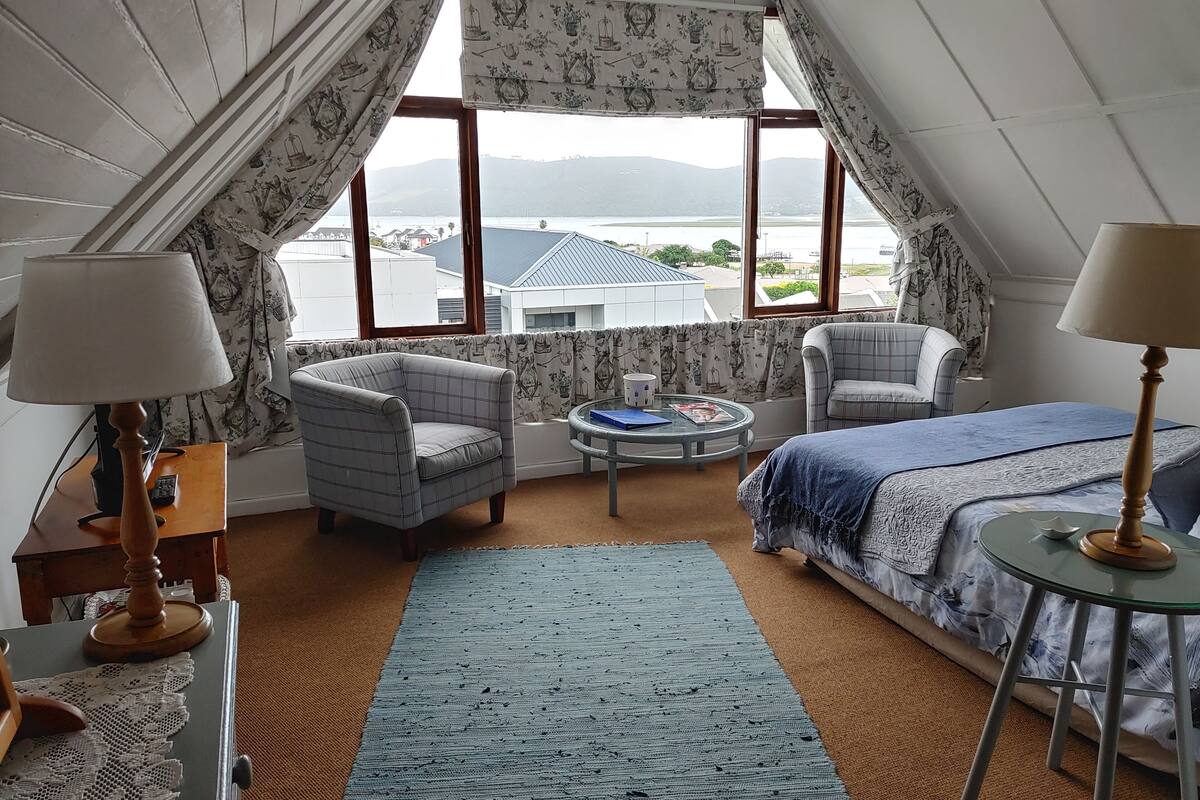 A cozy room with a bed, two armchairs, a small table, and a view of the sea through large windows.