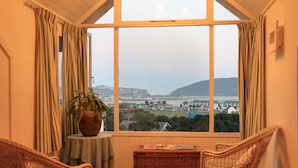 A room with a large window, wicker chairs, a table with a plant, and a view of a coastal town. - Yellowwood Lodge - Adults Only (Knysna)