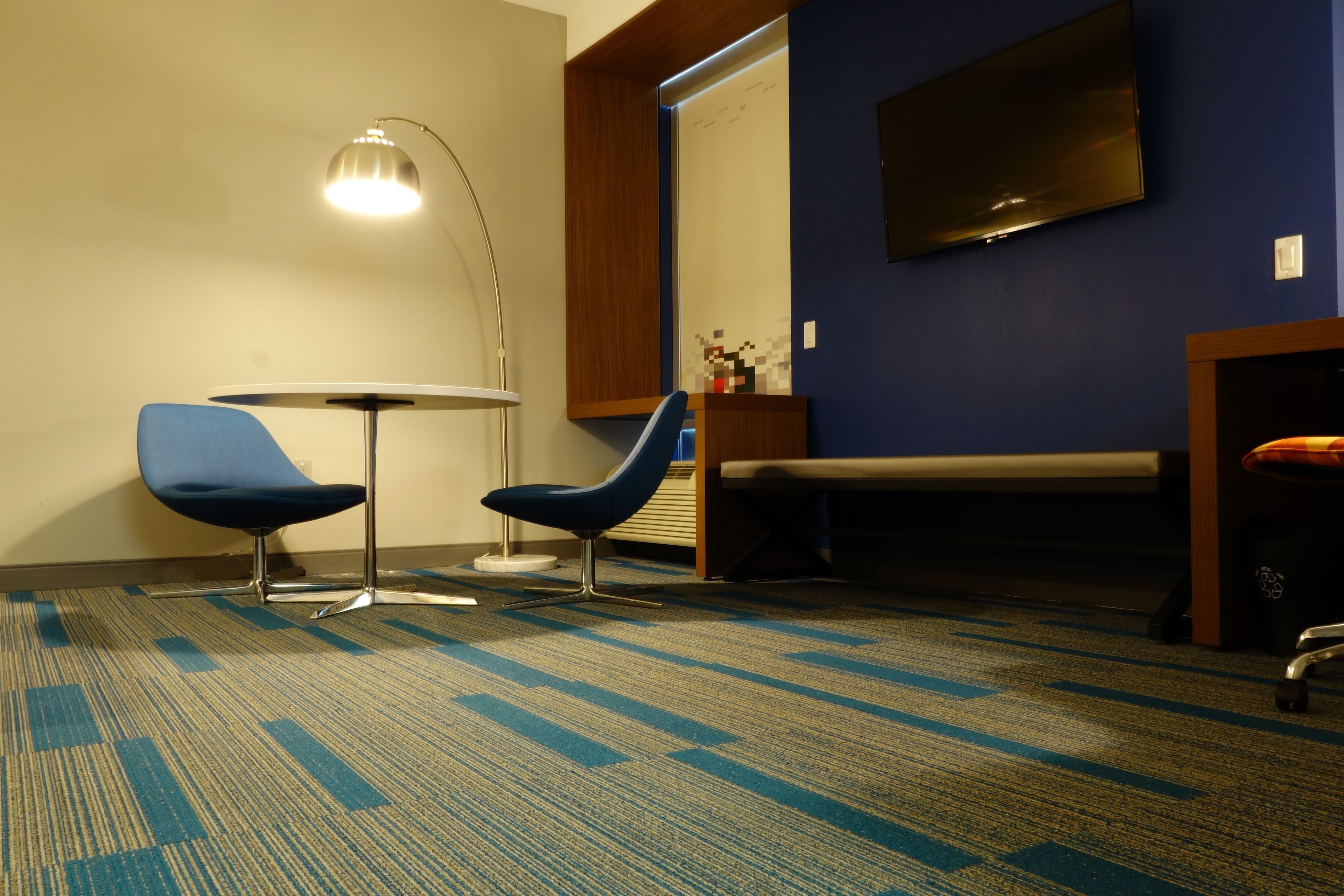 Habitación de hotel con alfombra azul, dos sillas azules, una mesa redonda, una lámpara de pie, un televisor en la pared y una cama con cabecero de madera.
