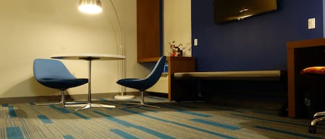 Habitación de hotel con alfombra azul, dos sillas azules, una mesa redonda, una lámpara de pie, un televisor en la pared y una cama con cabecero de madera.