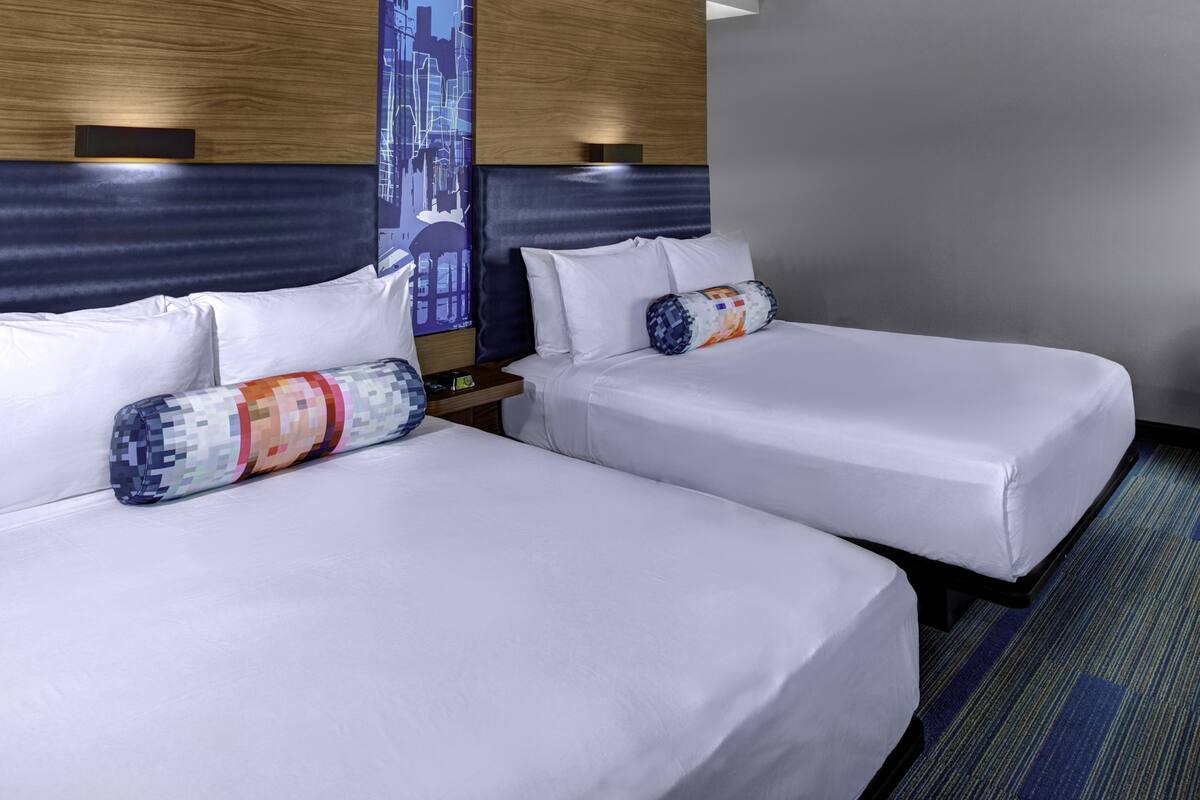 A hotel room with two single beds, each with white linens and a colorful pillow. There is a nightstand with a lamp between the beds. The room has a modern design with a wooden headboard and a cityscape mural on the wall.