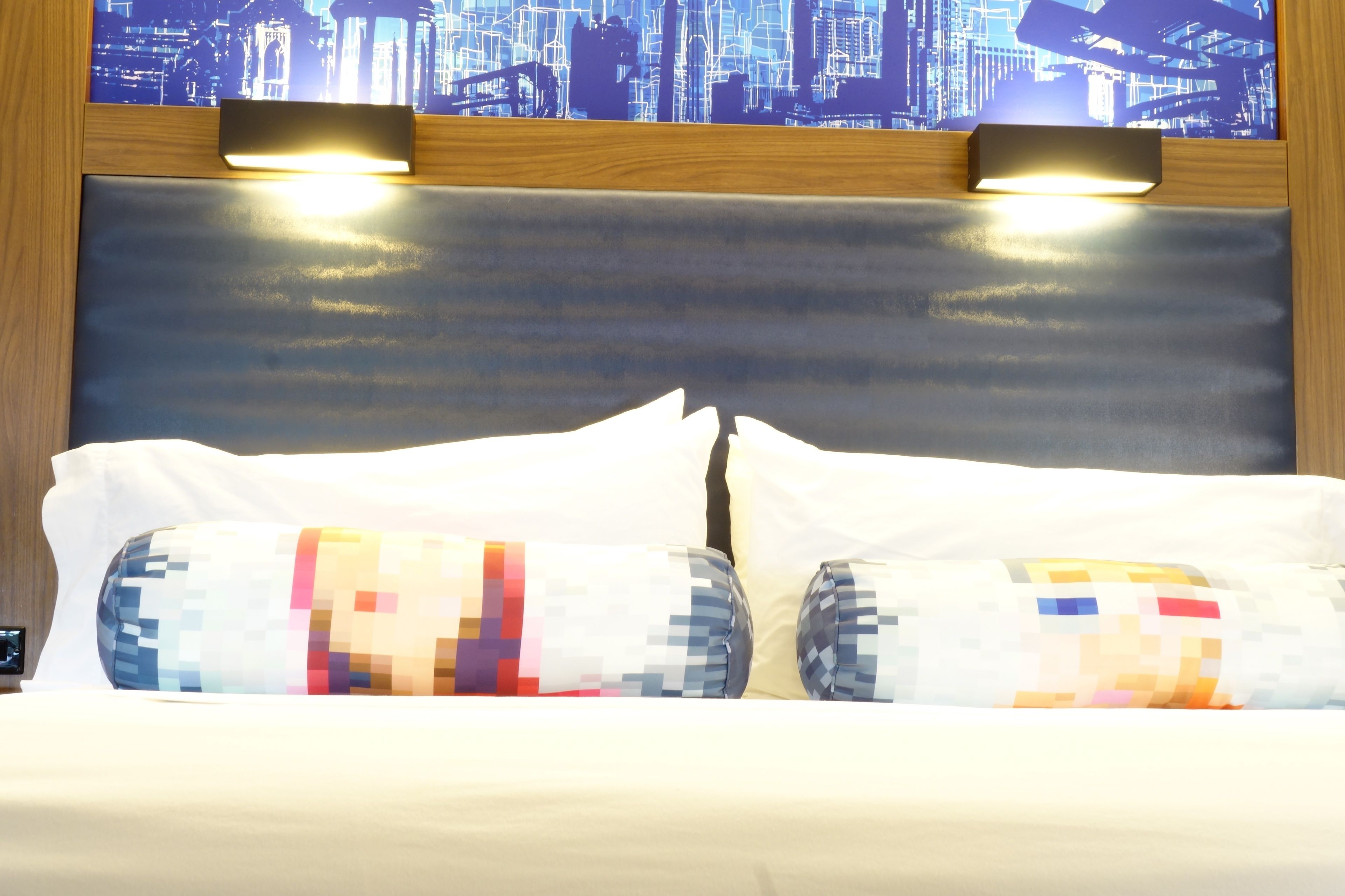 Ein Hotelzimmer mit einem großen Bett, zwei Kissen mit Pixelmustern, einem Holz-Kopfteil und Blick auf die Stadt.