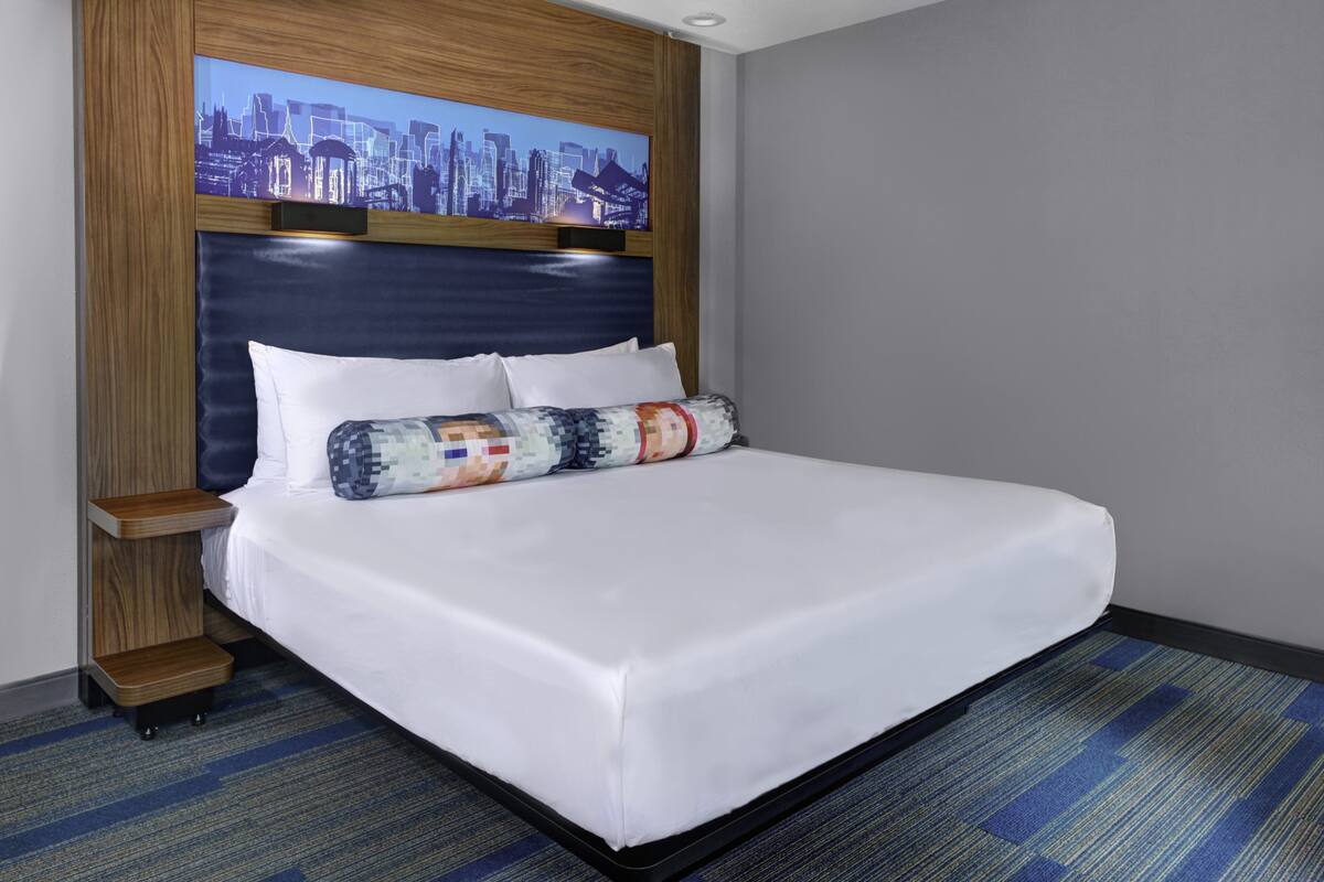 A hotel room with a large bed, a cityscape view, a wooden headboard, and a blue carpet.