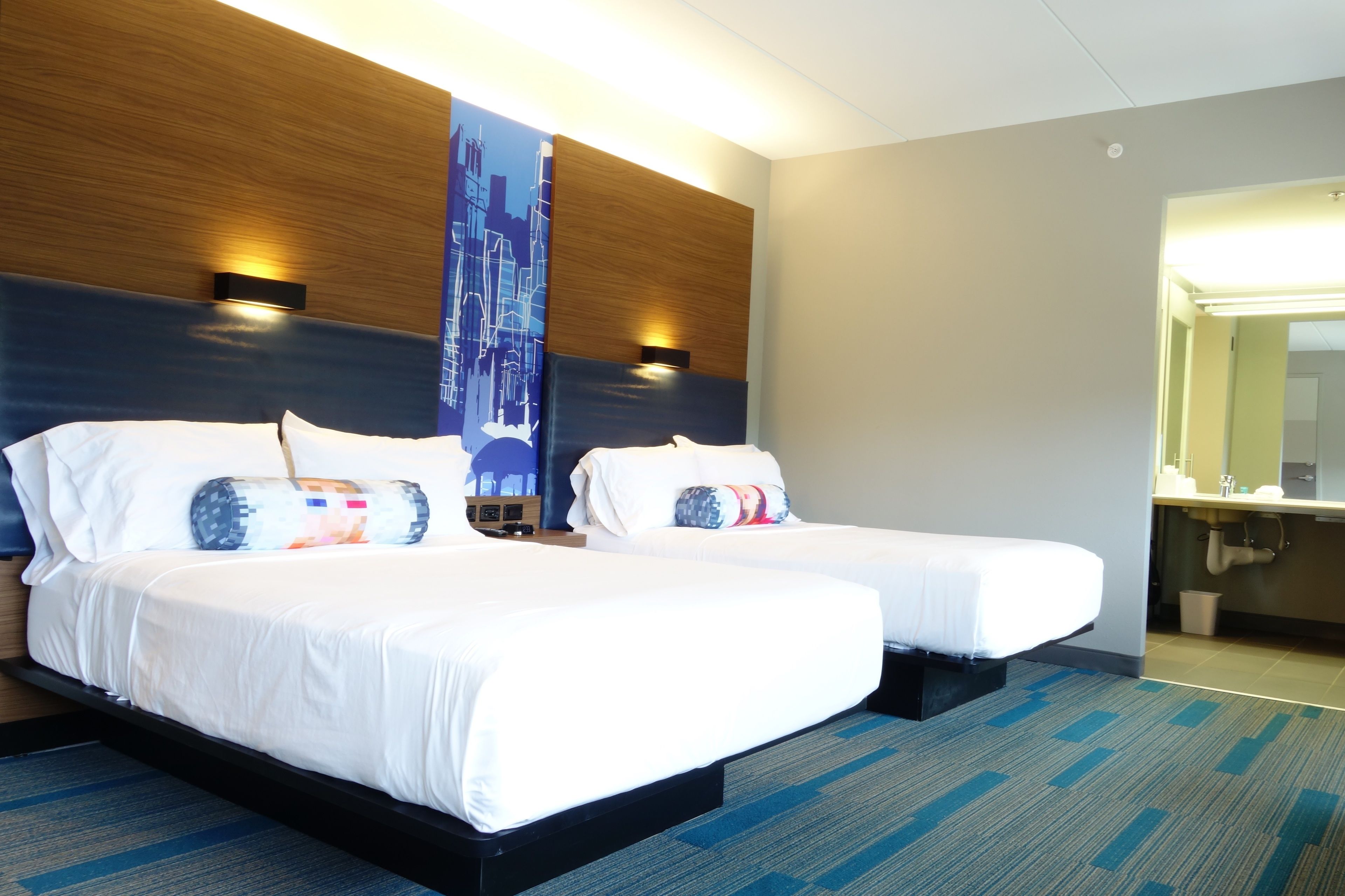 A hotel room with two beds, a large wall-mounted artwork, a wooden headboard, and a bathroom visible through an open door.