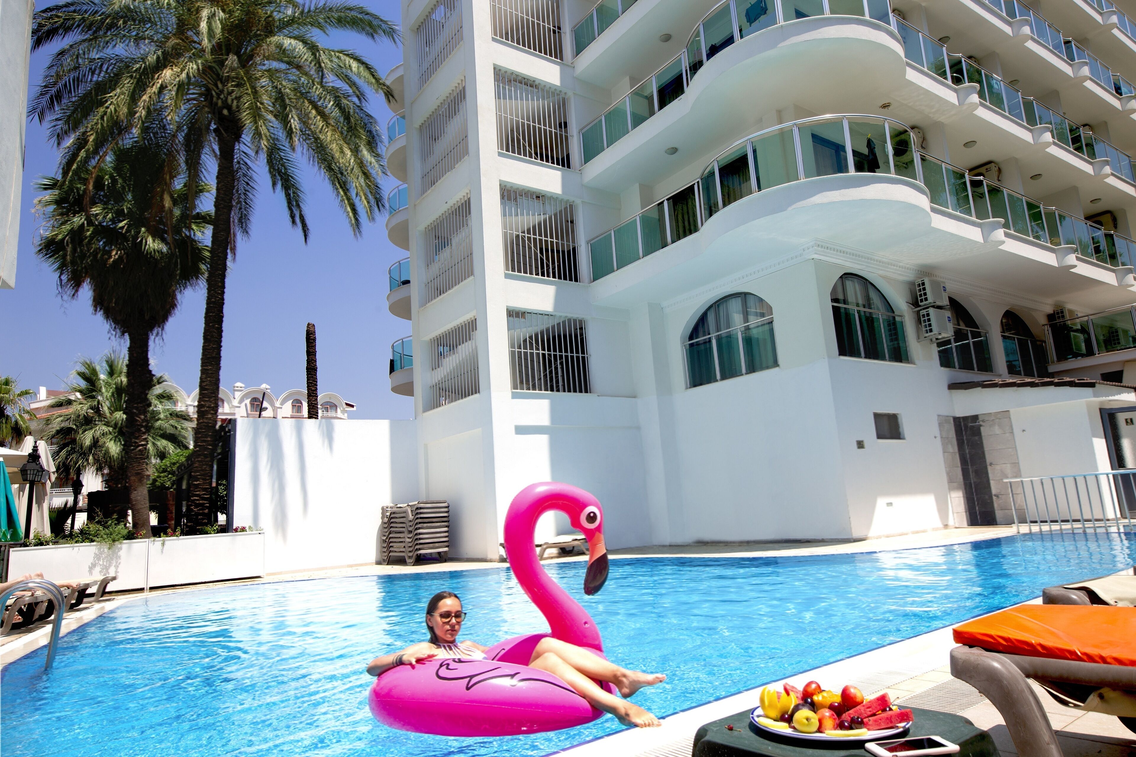 A swimming pool with a person on a pink flamingo float, palm trees, and a multi-story building with balconies.