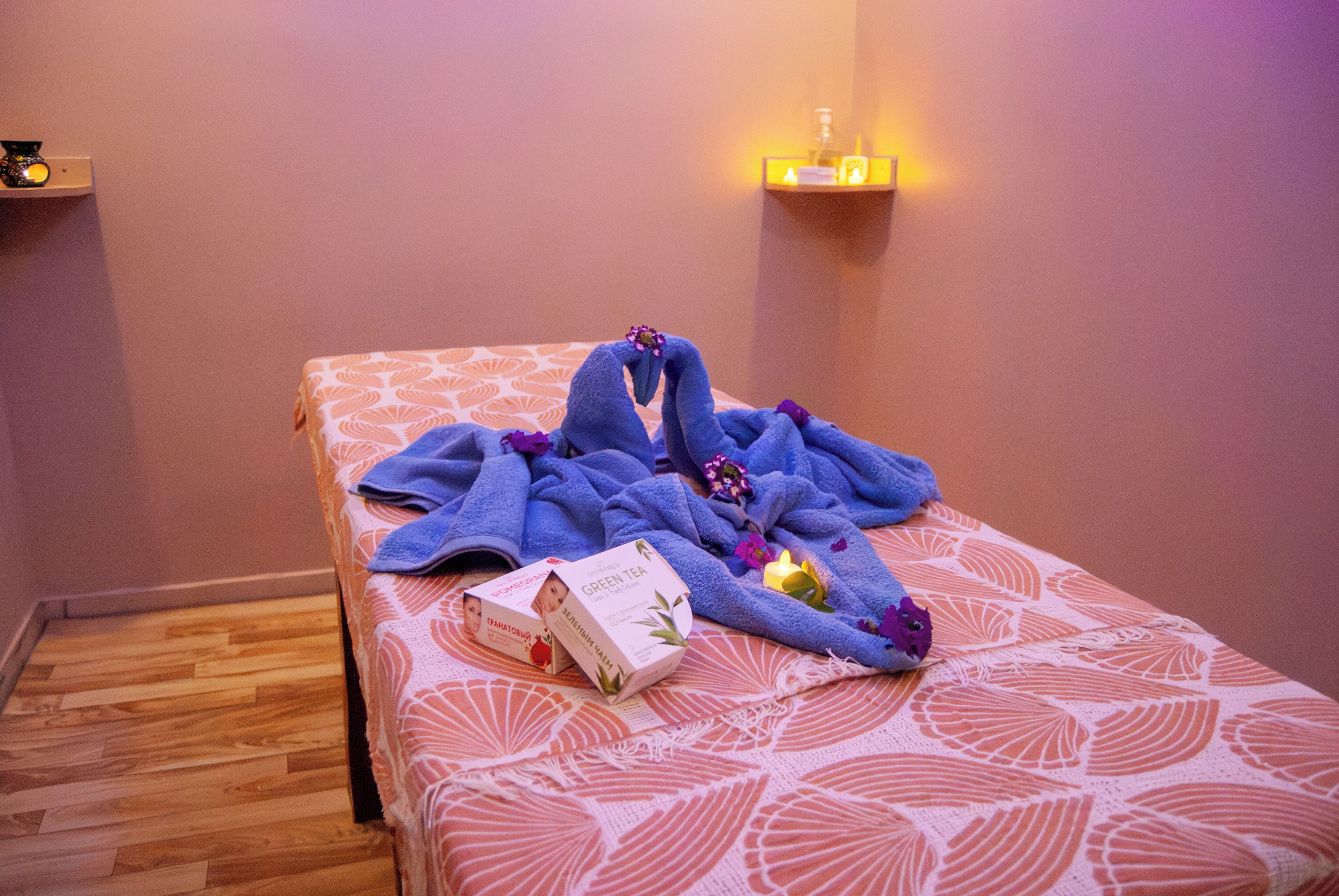 A neatly made bed with a patterned bedspread, a blue towel, and a box of green tea.